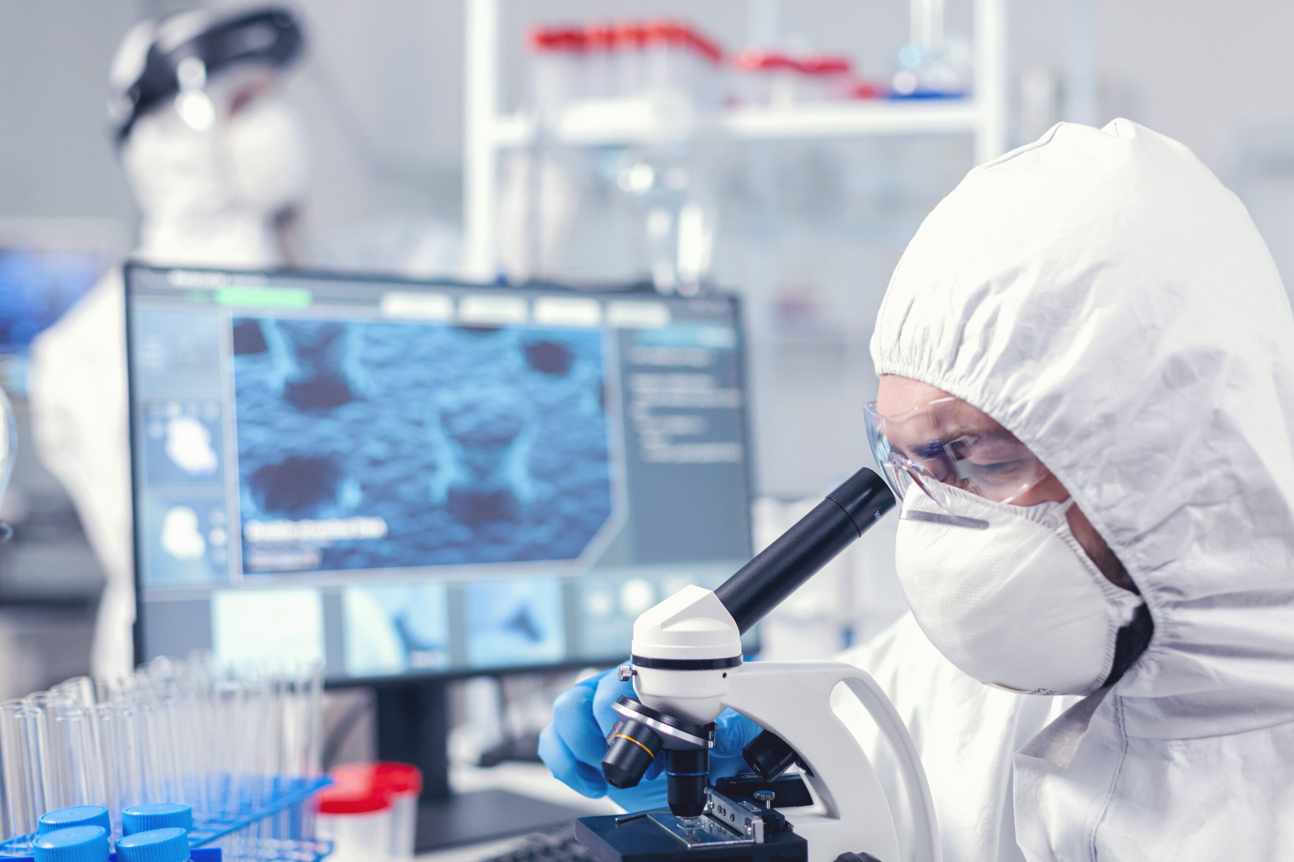 Medical team in laboratory conducting coronavirus analysis using microscope. Scientist in protective suit sitting at workplace using modern medical technology during global epidemic.